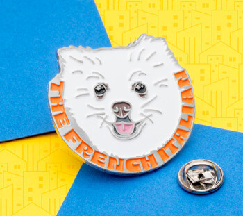 Enamel lapel pin featuring a white fluffy dog face with orange text reading The French Italian.