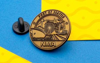 Circular antiqued gold lapel pin featuring the Port of Seattle skyline with a ship, Ferris wheel, and airplane above the text NMA.