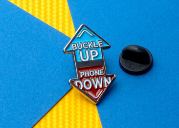 Double-sided arrow lapel pin with 'Buckle Up' in blue and 'Phone Down' in red soft enamel.