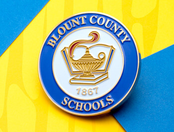 Circular lapel pin for Blount County Schools featuring a gold lamp of knowledge and book icon on a blue and white background.