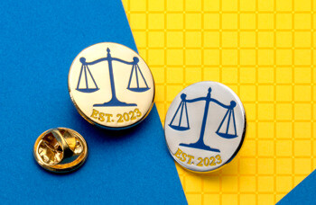 Gold and silver lapel pins featuring blue scales of justice and Est. 2023 text on a blue and yellow background.