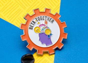 Beta Together lapel pin featuring an orange gear shape with a purple silhouette head and colorful icons including a lightbulb and paintbrush.
